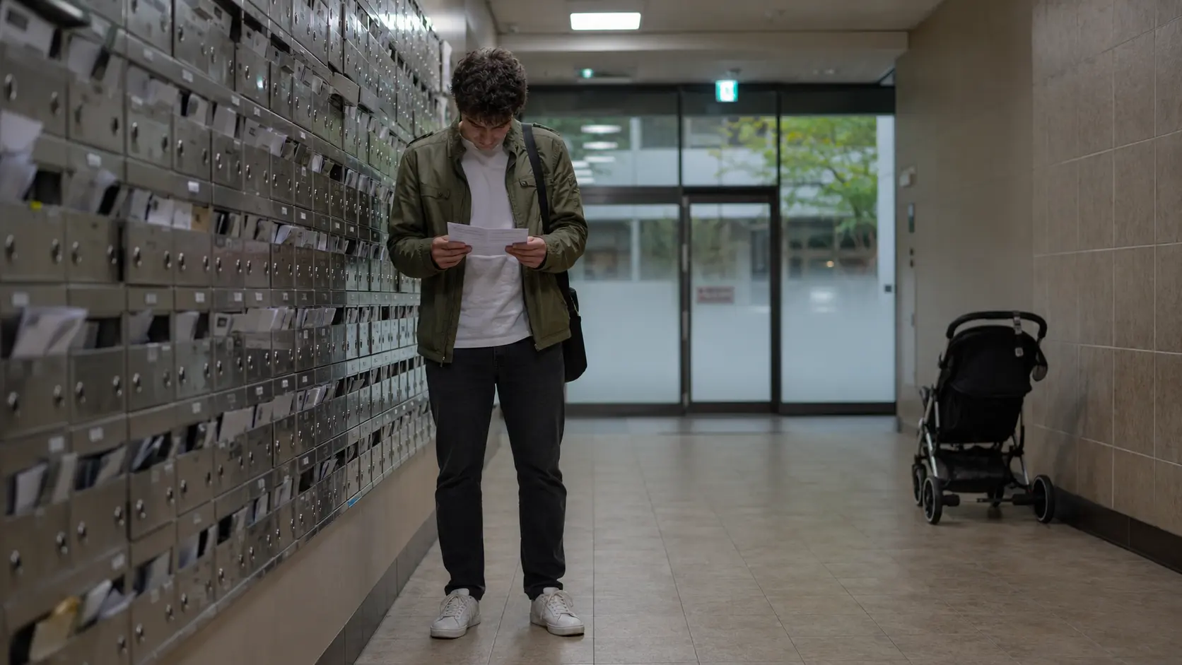 Foreign resident in olive-green zip-up jacket and dark grey jeans reading a Korean apartment maintenance fee bill in a mailbox lobby