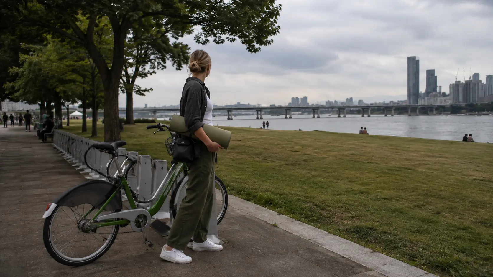 Foreign visitor carrying a picnic mat beside a rental bike on a Han River path